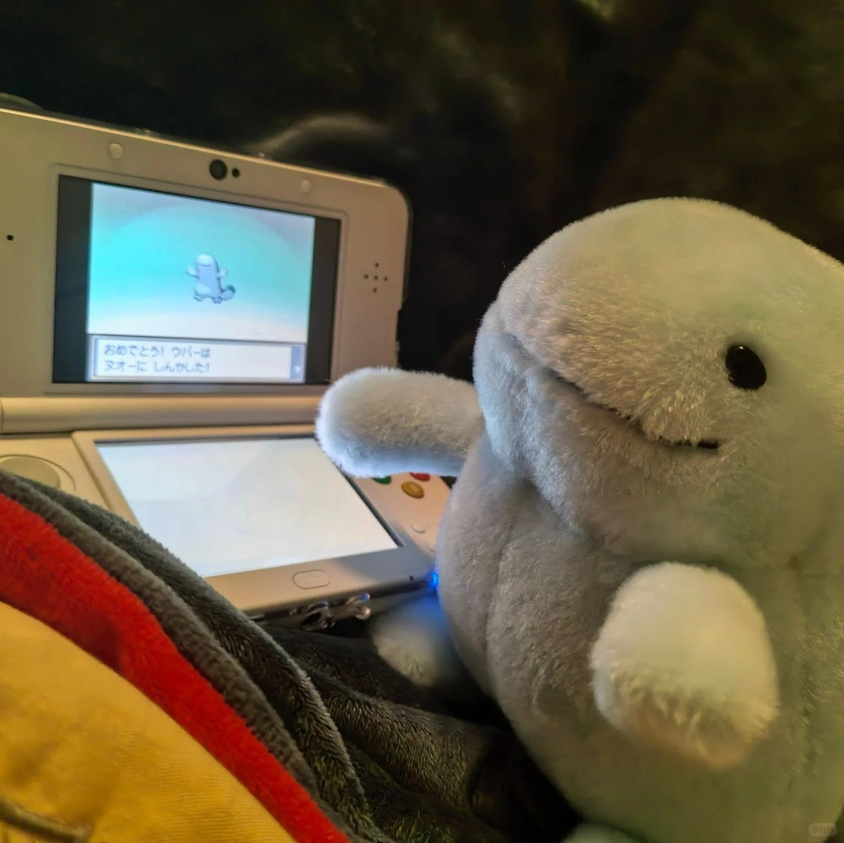 A light blue  Quagsire  plush toy with a stitched smile sits next to a white handheld gaming console on a dark couch, with a red and yellow blanket in the foreground.