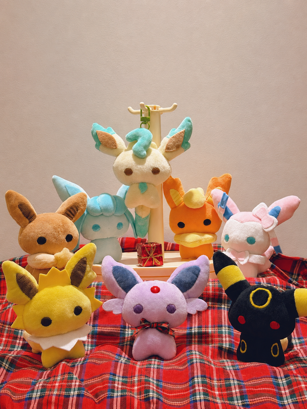 A collection of plush toy characters arranged on a red plaid blanket against a plain beige wall. The plushies include Eevee, Vaporeon, Flareon, Eevee, Eevee, Eevee, and Umbreon, with one standing on a small display stand.