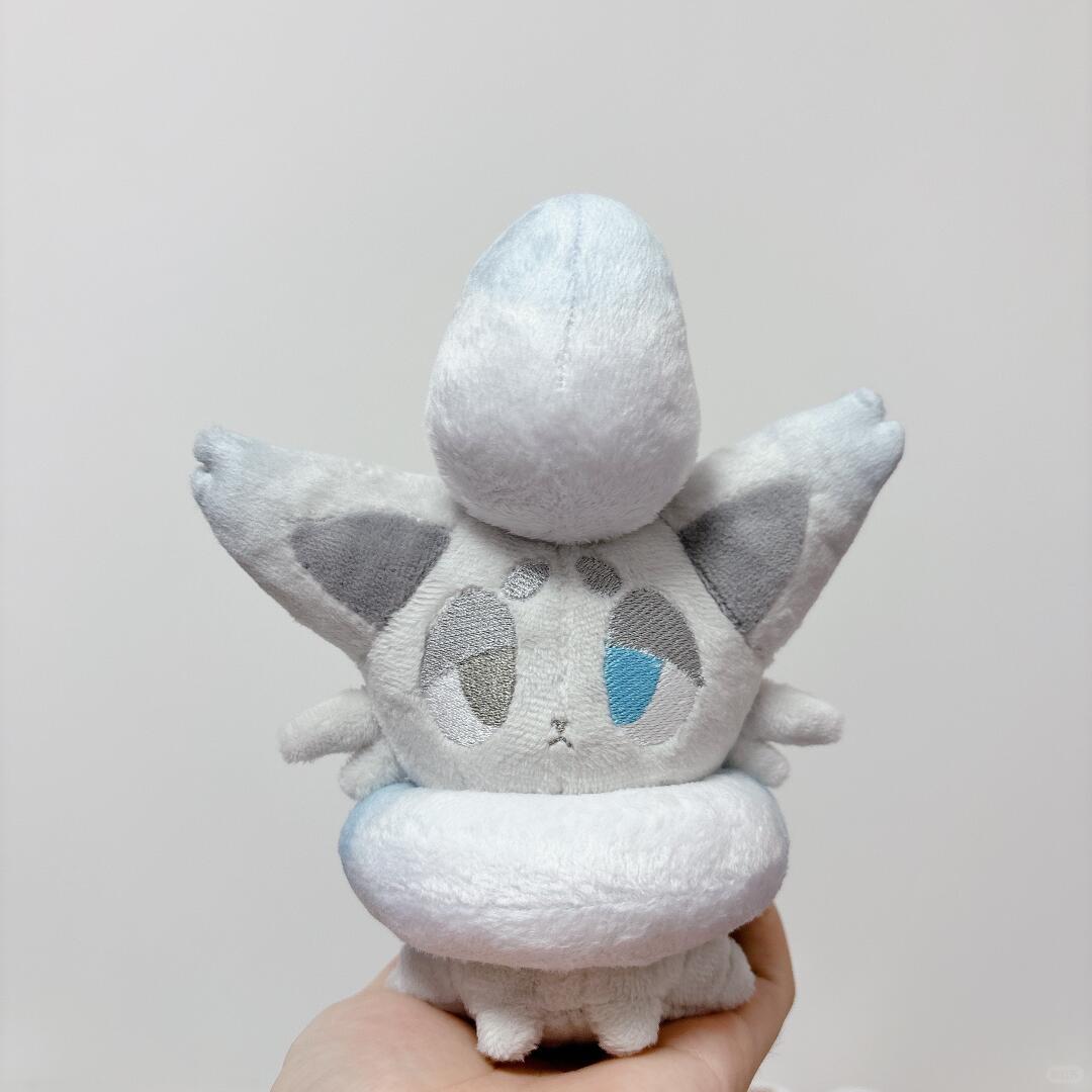 A plush toy of a Zorua white and gray winged creature with a blue teardrop detail on its face, held in a person's hand against a plain light gray background.
