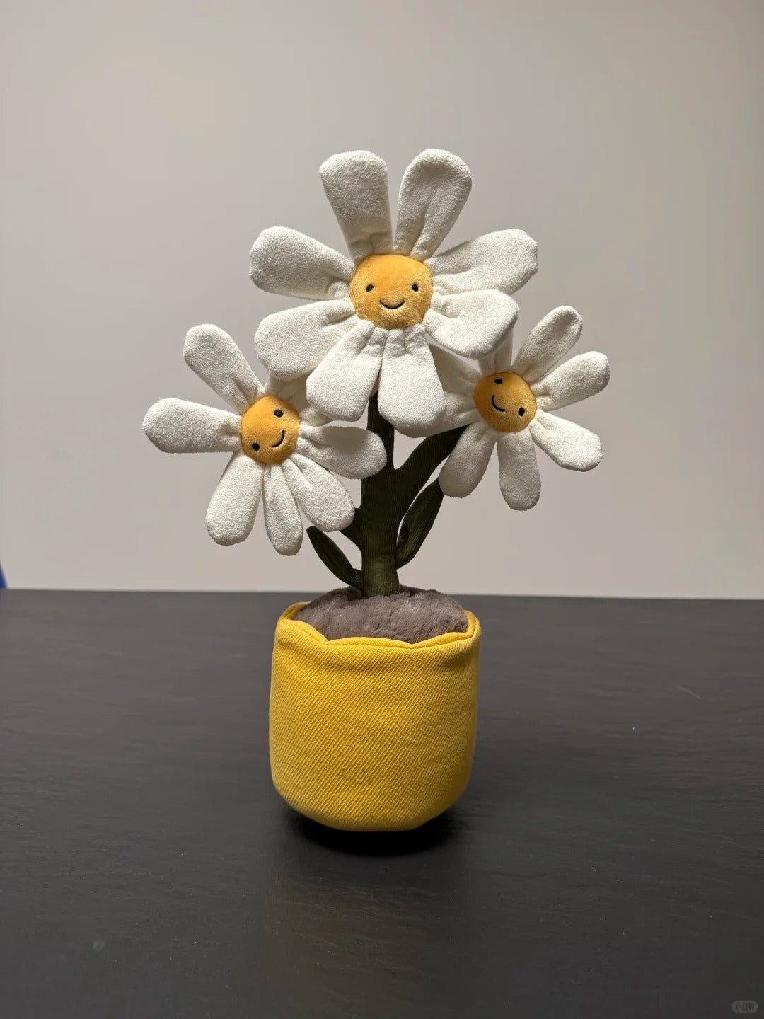 A handmade plush toy featuring three smiling white daisy flowers with yellow centers, planted in a yellow fabric pot on a dark tabletop against a plain light gray wall.