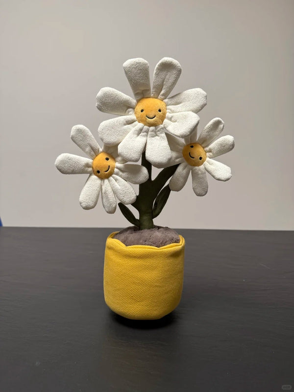 A handmade plush toy featuring three smiling white daisy flowers with yellow centers, planted in a yellow fabric pot on a dark tabletop against a plain light gray wall.