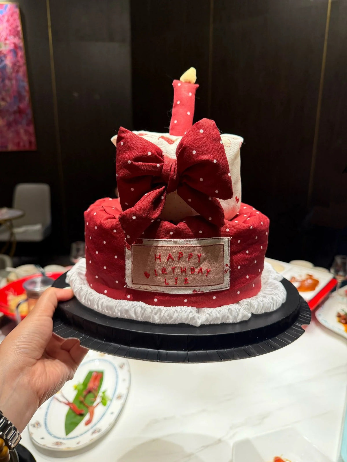 A hand holds a two-tiered red and white polka dot cake decorated with a large red bow, a candle, and a 'HAPPY BIRTHDAY LEE' plaque, set against a dark restaurant background.