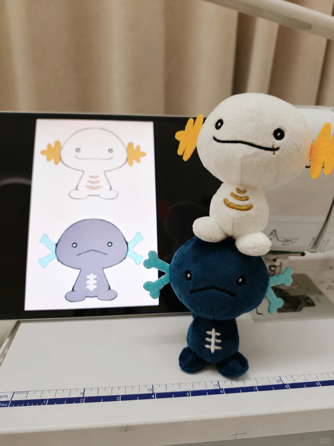 Two Wooper plush axolotl toys displayed on a white table in front of a laptop showing axolotl design sketches. One plush is white with yellow gills and a gold band, sitting on top of a dark blue plush with teal gills and white belly details.