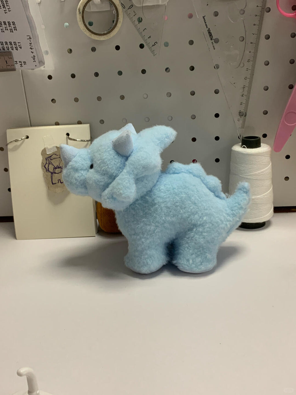 Blue plush dinosaur toy on a white surface with a pegboard background