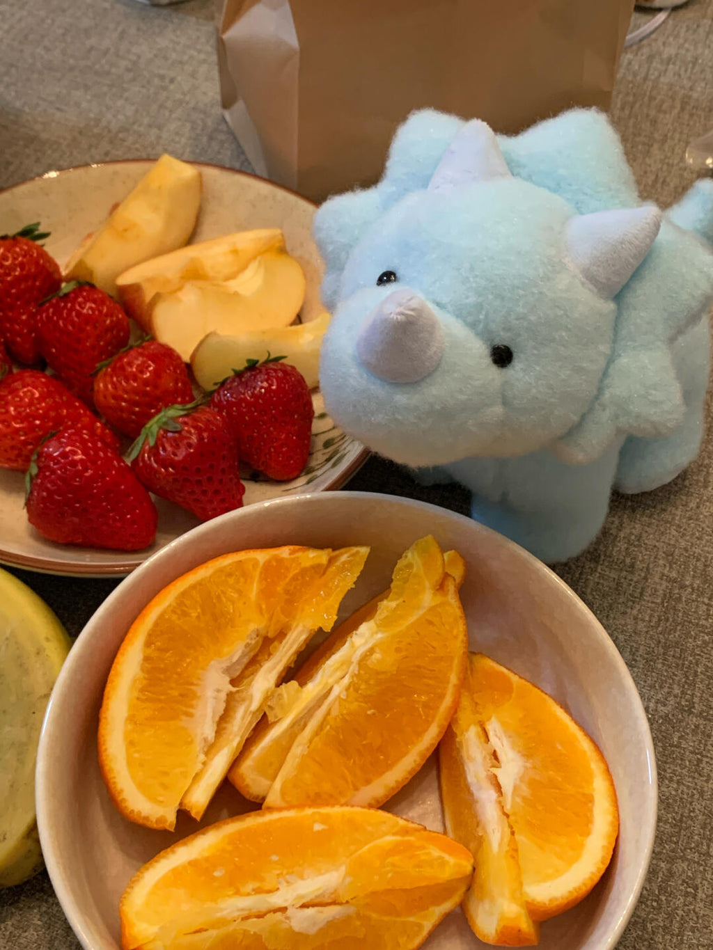 Blue dinosaur plush toy next to a bowl of orange slices and a plate of fruit on a carpeted floor.