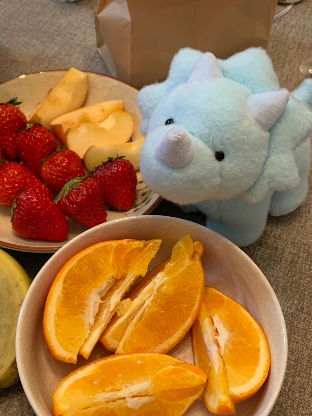 Blue dinosaur plush toy next to a bowl of orange slices and a plate of fruit on a carpeted floor.
