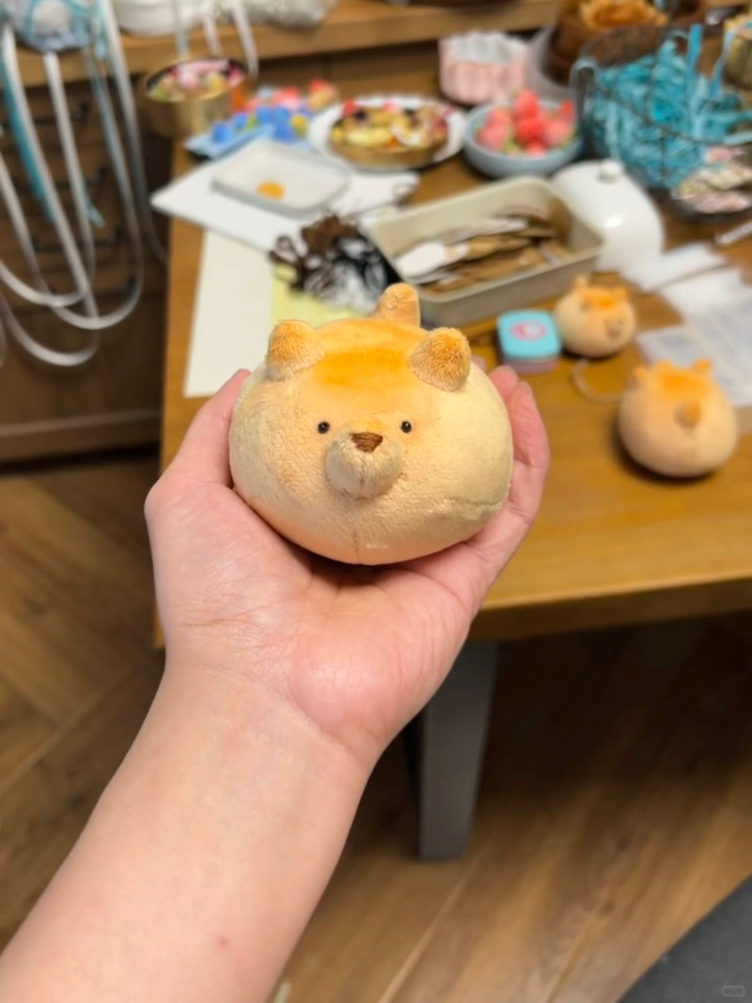 A hand holding a small plush toy shaped like a round, soft-faced character with short ears and embroidered facial features, with a wooden table and other plush items in the background.
