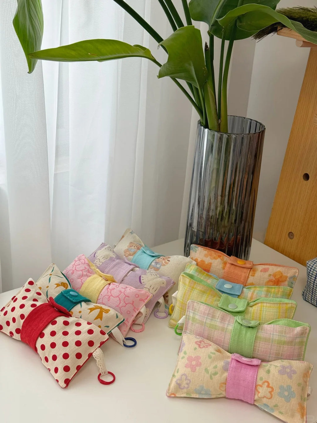 A group of small fabric glass pouches or sachets in assorted prints and patterns, tied with ribbon and small plastic rings, arranged on a white table next to a tall glass vase with green leaves.