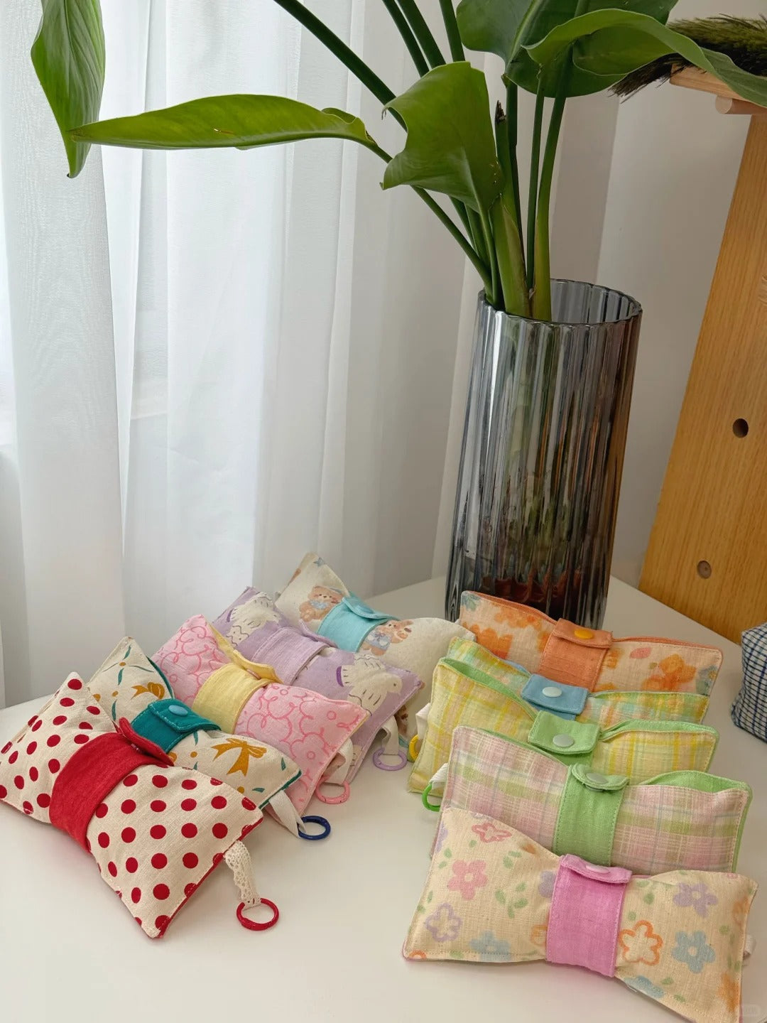A group of small fabric glass pouches or sachets in assorted prints and patterns, tied with ribbon and small plastic rings, arranged on a white table next to a tall glass vase with green leaves.