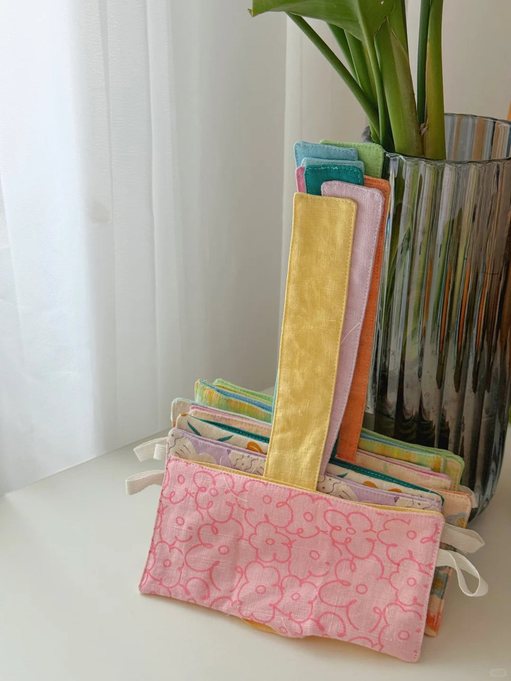 A stack of colorful fabric glass pouches or small zippered cases with long handles, displayed on a white surface next to a tall ribbed glass vase with green leaves. The front pouch is pink with a lighter pink floral pattern.