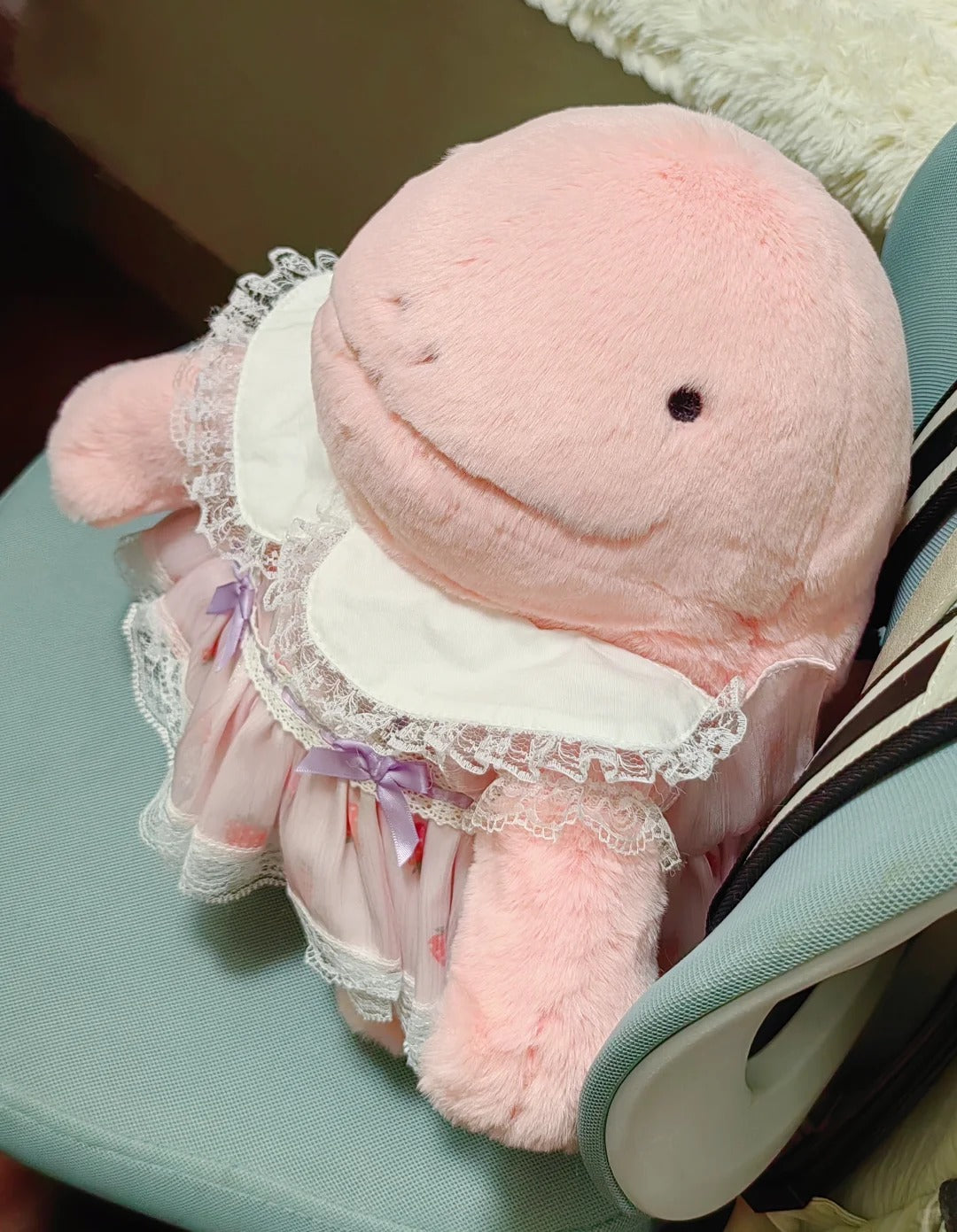 A pink Quagsire plush whale toy wearing a white bib and a pink ruffled dress with lace trim and a small purple bow, resting on a light blue chair.