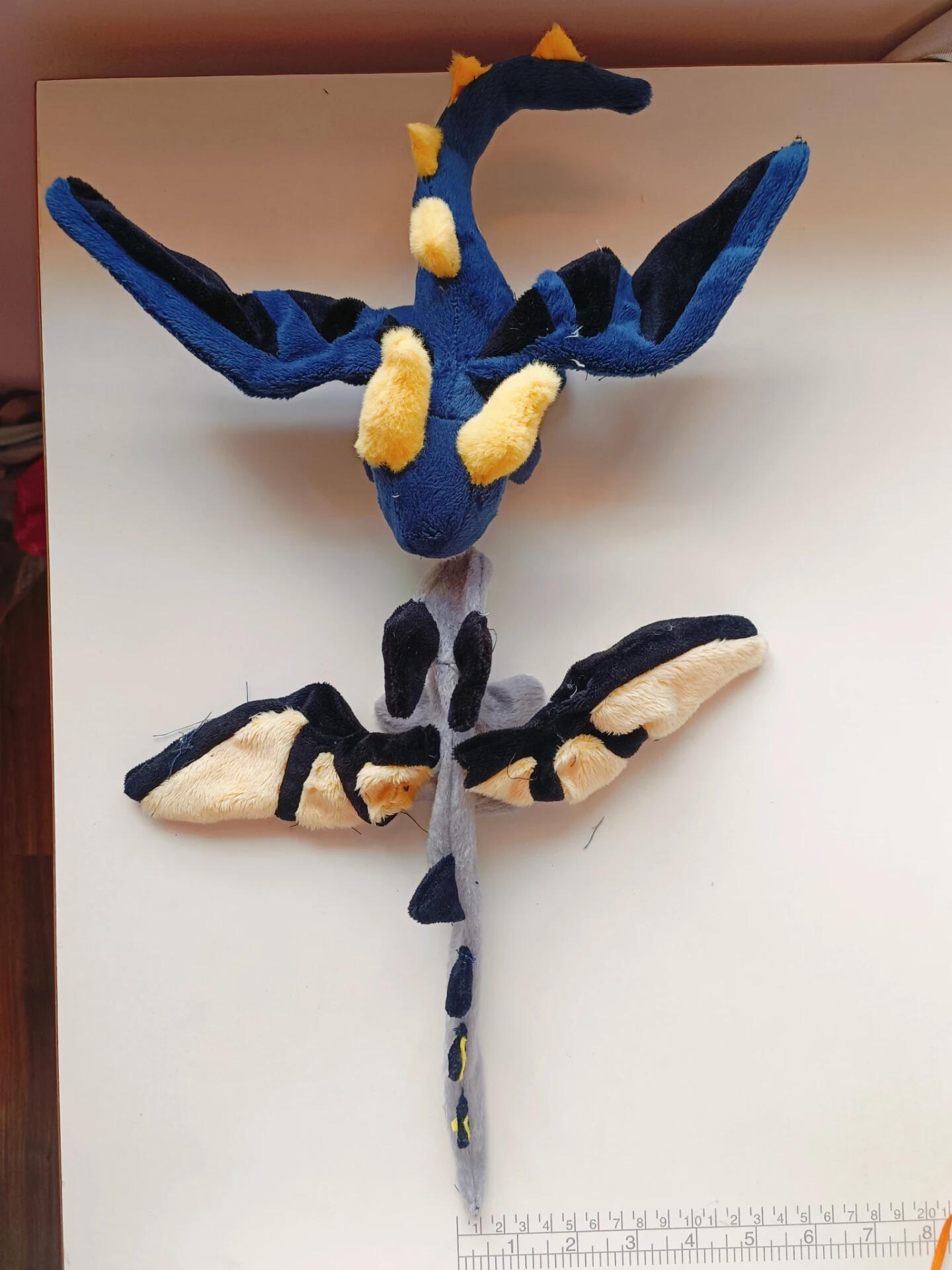 Plush toy of a blue and yellow creature on a white surface with a ruler for scale.
