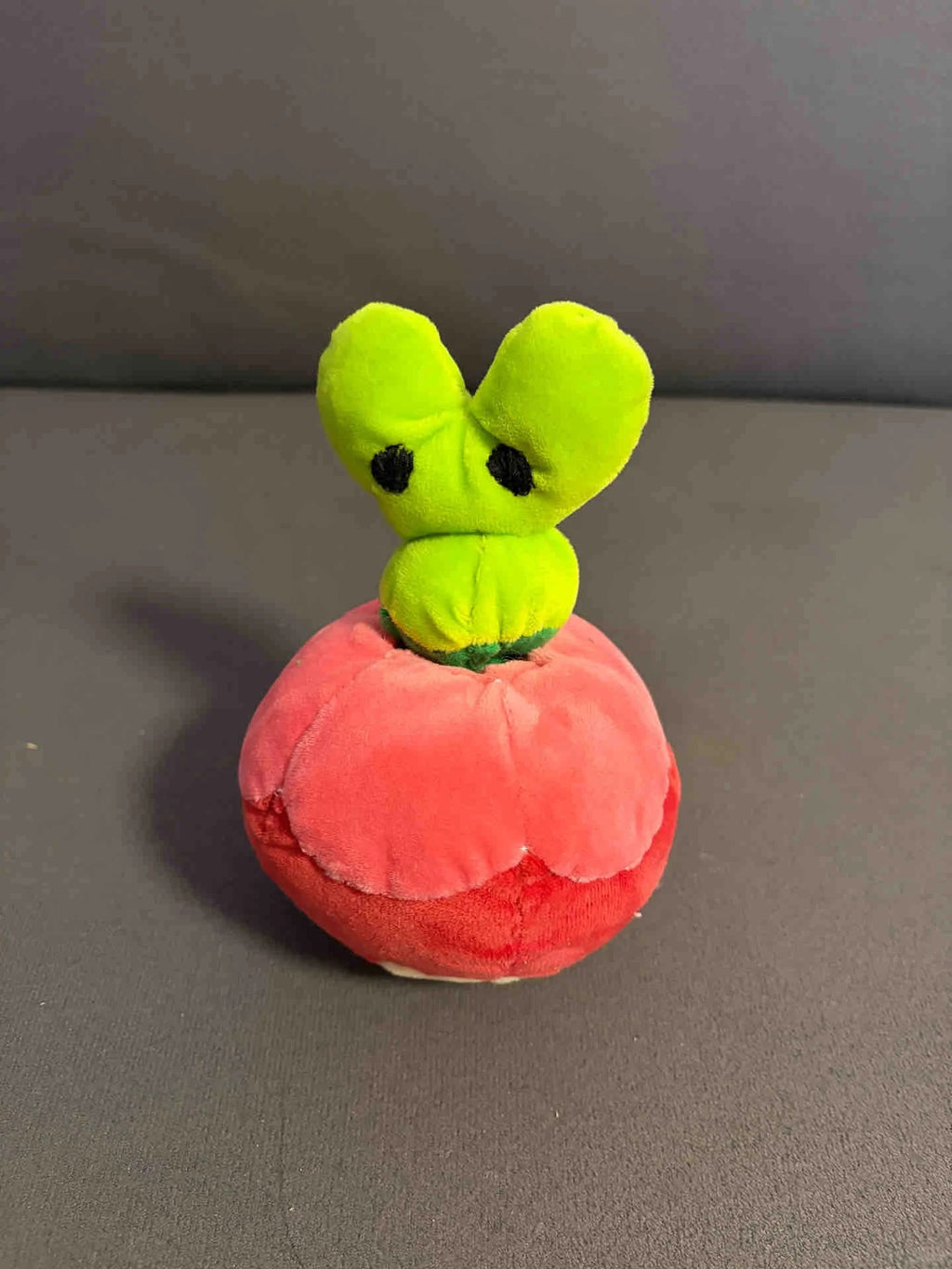 A Applin plush toy shaped like a tomato with a bright green leafy top and black eyes, sitting on a red tomato-shaped base against a gray background.