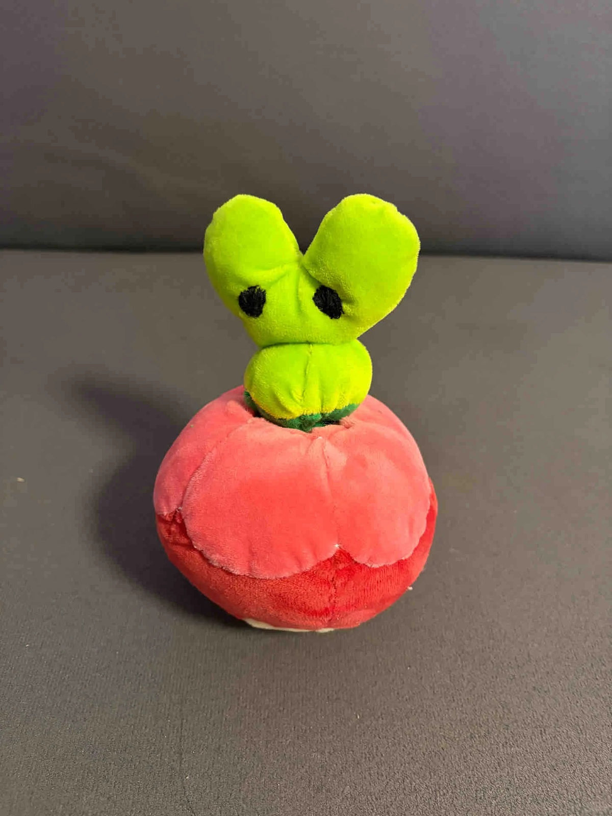 A Applin plush toy shaped like a tomato with a bright green leafy top and black eyes, sitting on a red tomato-shaped base against a gray background.