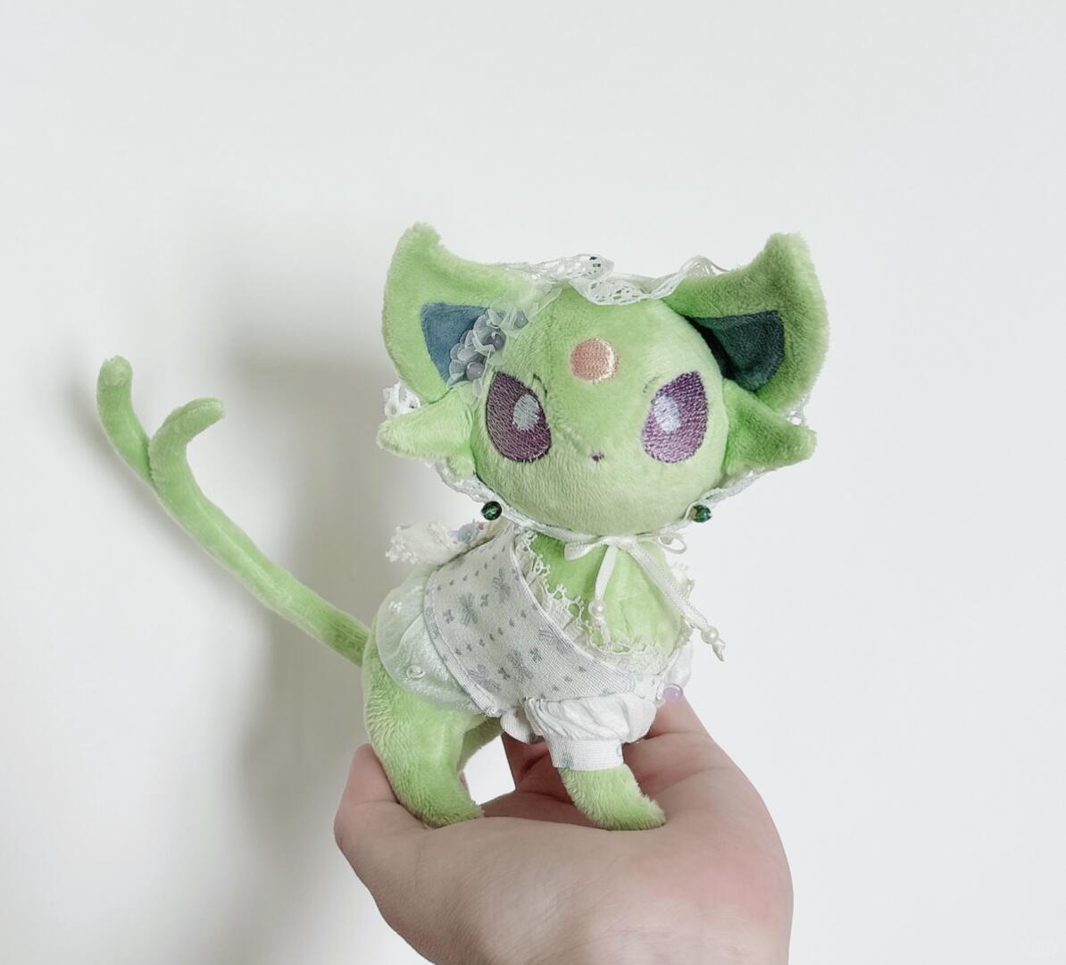 A small green pokemon eevee plush toy with large pointed ears, purple eyes, and a white lace dress, being held in a person's hand against a plain light background.