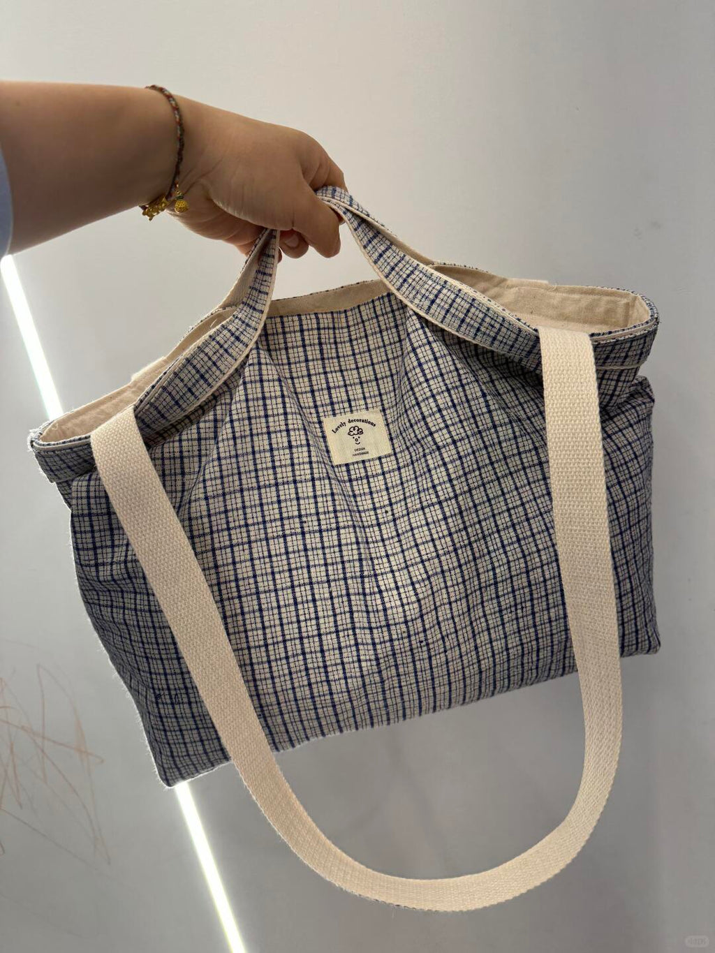 A hand holding a blue and white checkered canvas tote bag with thick cream-colored shoulder straps against a plain light gray wall.