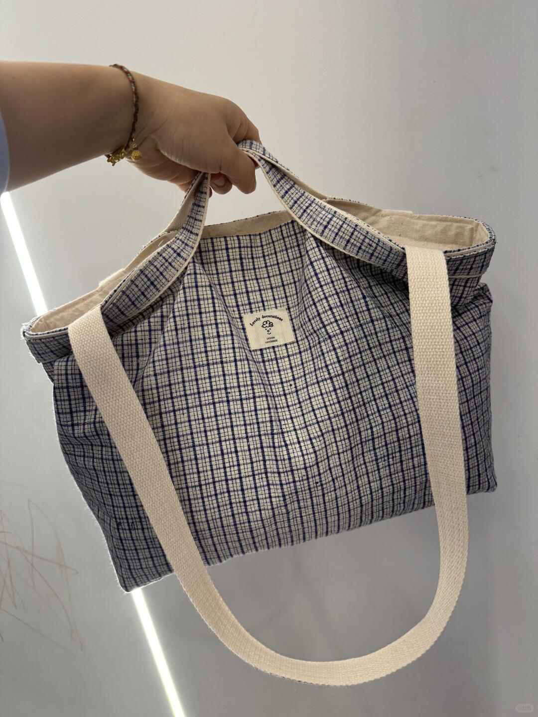 A hand holding a blue and white checkered canvas tote bag with thick cream-colored shoulder straps against a plain light gray wall.