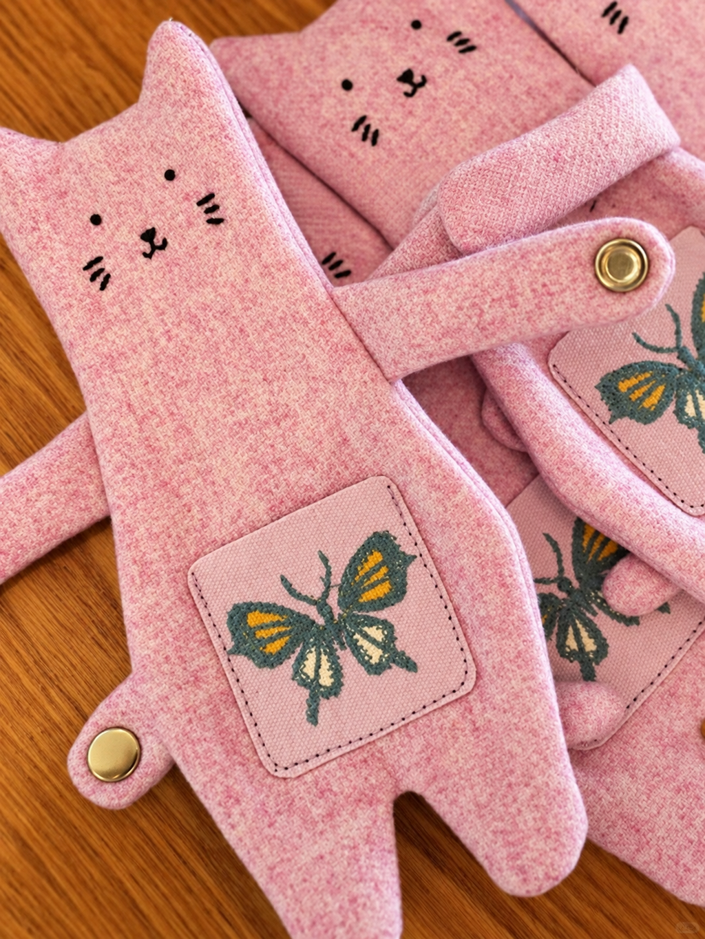 Pink felt cat-shaped gloves with embroidered faces and butterfly patches, arranged on a wooden surface.