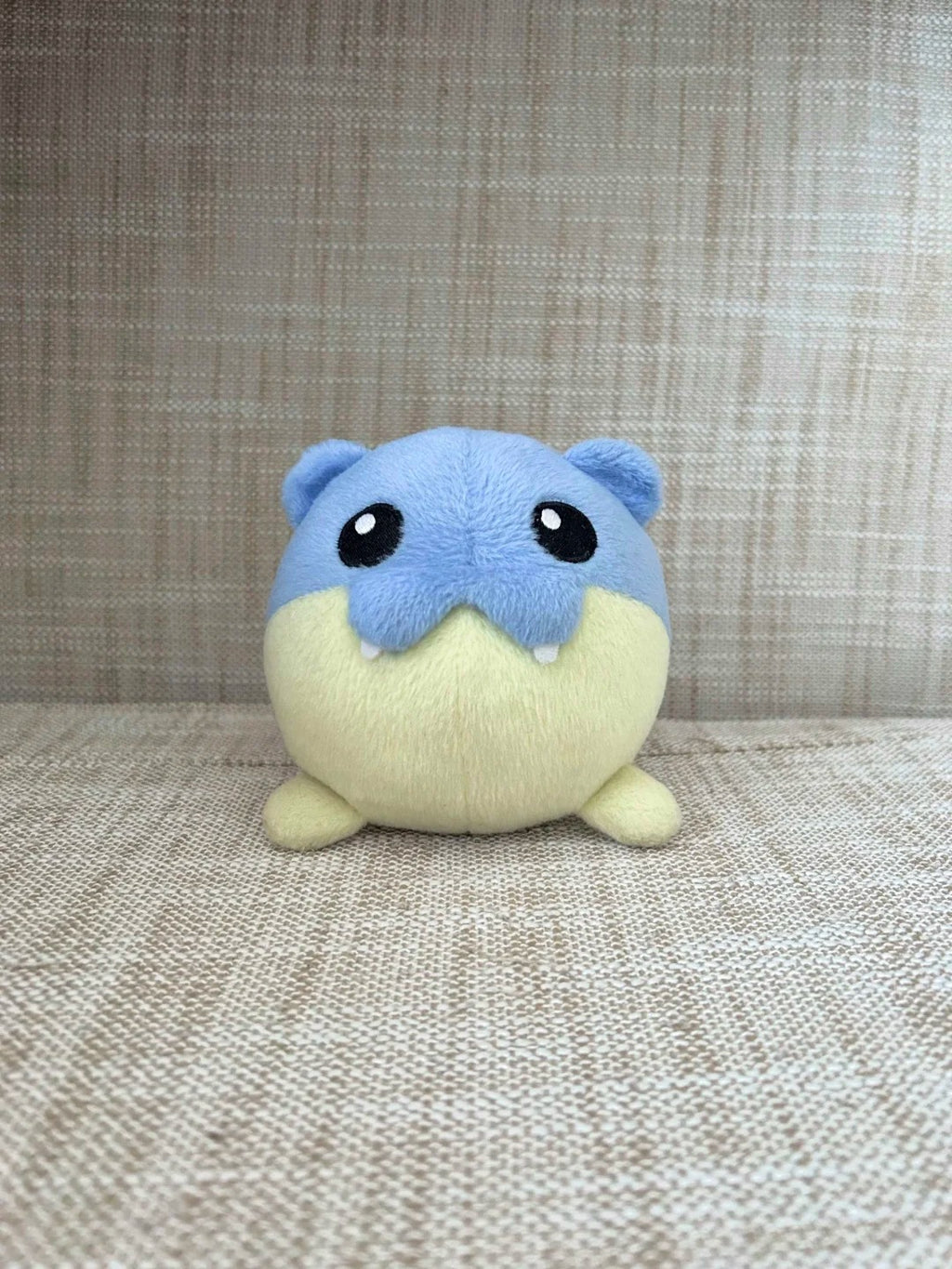 A small Spheal plush toy resembling a blue and cream-colored aquatic creature, photographed against a textured beige woven fabric background.
