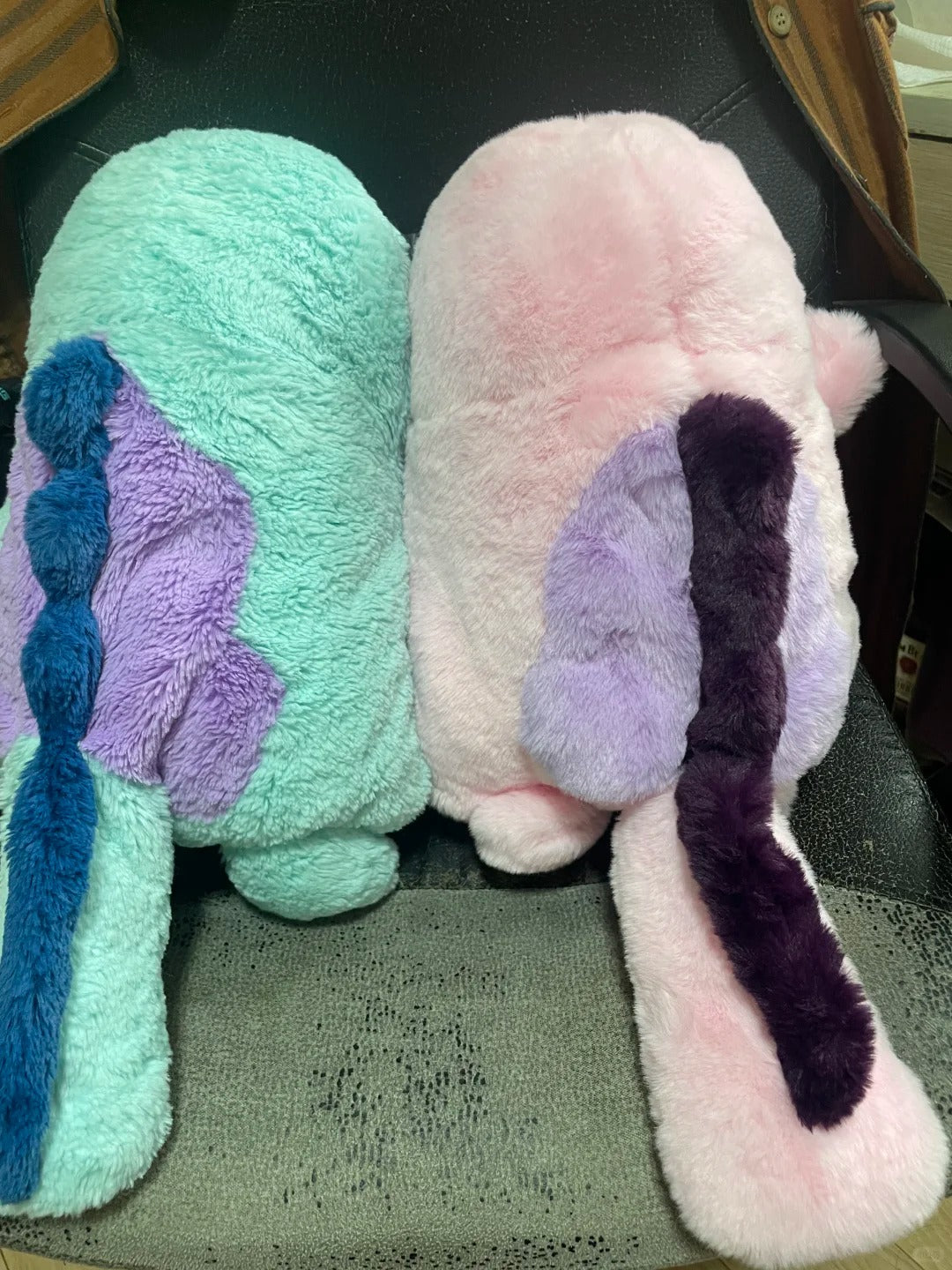 Two plush Quagsire  sitting on a gray carpeted floor. The left one is mint green with a purple wing and blue tail, and the right one is light pink with a purple wing and dark purple tail.