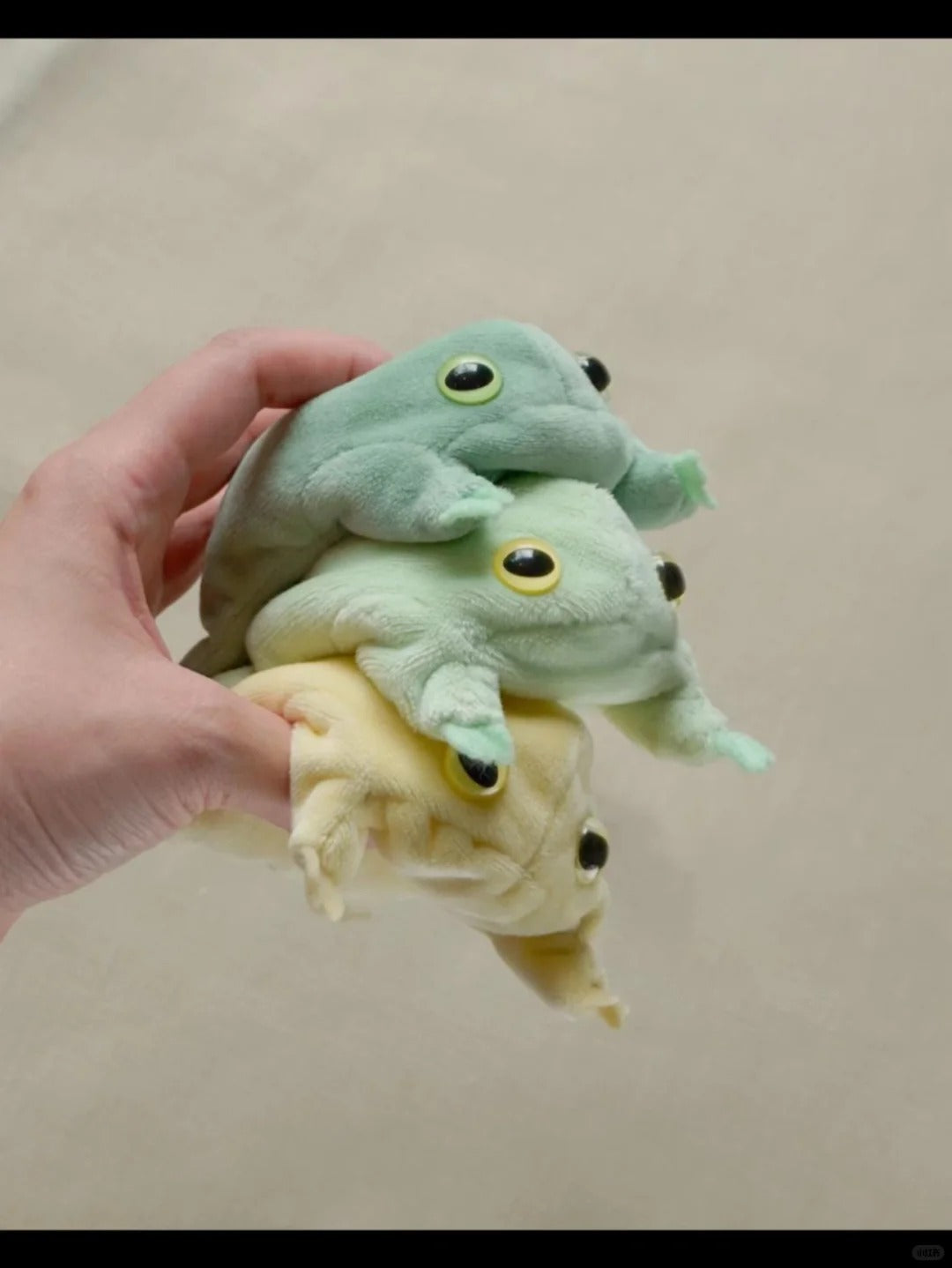 Three small frog plush toys in mint green, pale green and yellow stacked in one hand.