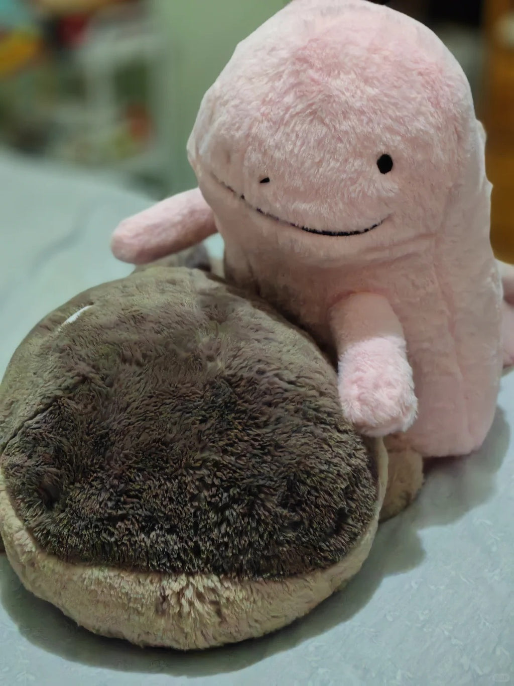 A  Quagsire  pink plush toy with a simple stitched smile and small black eyes, posed behind a round brown and beige plush cushion on a light surface.