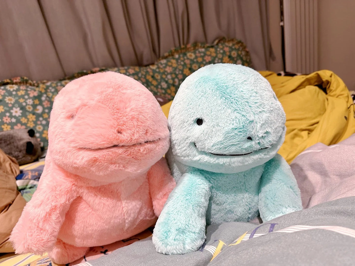 Two plush Quagsire dinosaur toys sitting side by side on a bed. The left plush is light pink, and the right plush is light blue.