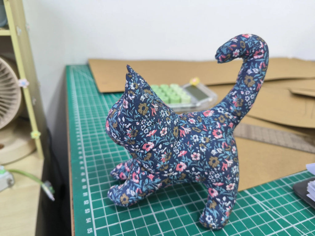 A small fabric cat figurine with a blue floral print, standing on a green cutting mat in a workshop setting.