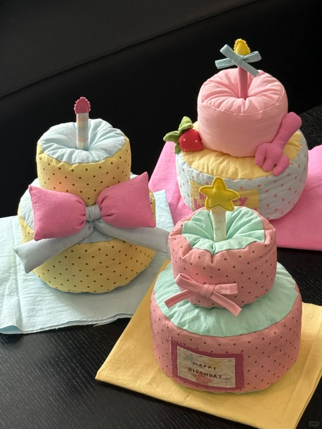 Three plush diaper cake toys arranged on a dark tabletop with napkins. The cakes are made from folded diapers and decorated with bows, polka dots, stars, candles, and small fruit accents in pink, yellow, mint green, and white.