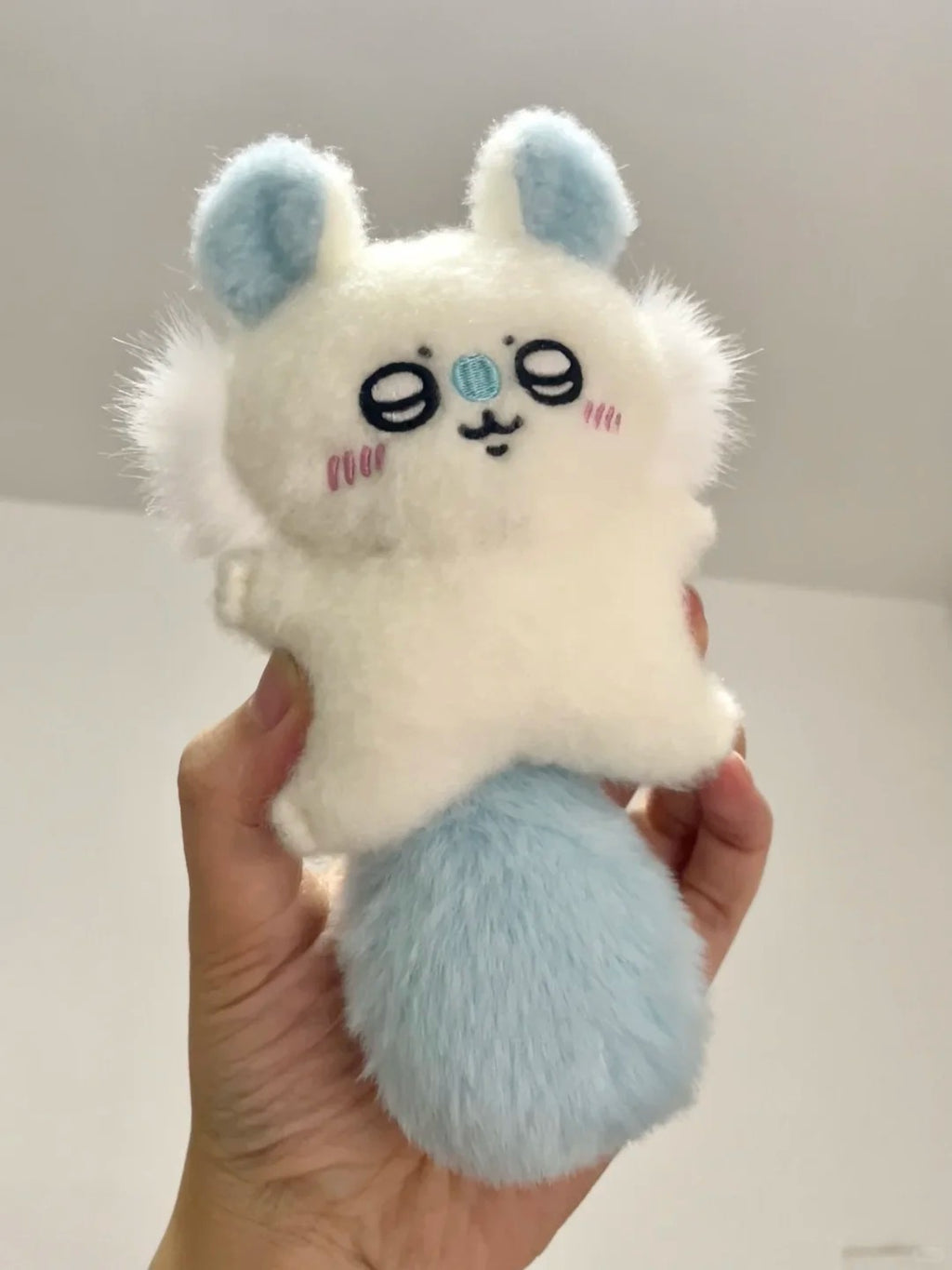 A small white momonga plush character toy with light blue ears and tail, held in a person's hand against a plain indoor background.