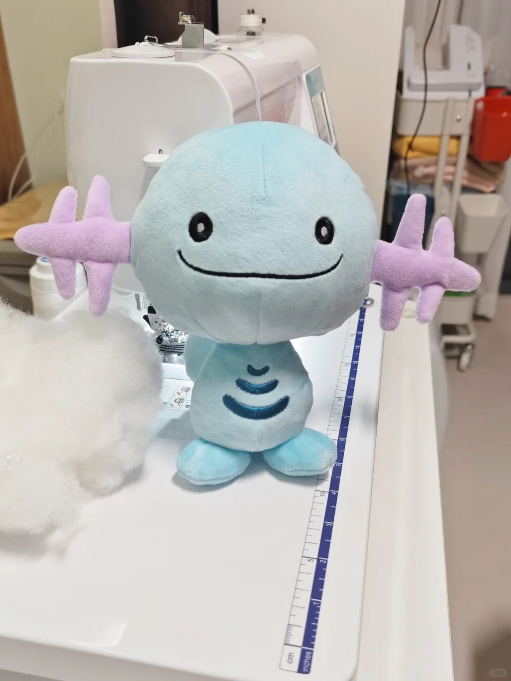 A light blue and lavender Wooper plush toy with a round head, black embroidered eyes and smile, and star-shaped arms, displayed on a white sewing machine table with a measuring tape nearby.