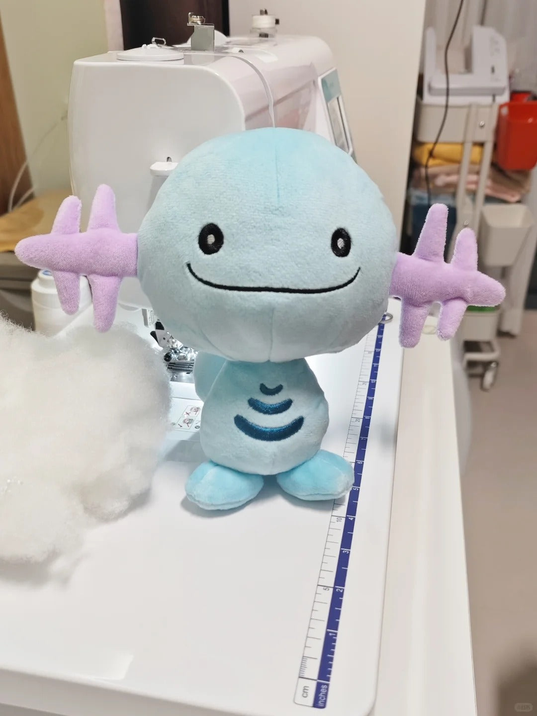 A light blue and lavender Wooper plush toy with a round head, black embroidered eyes and smile, and star-shaped arms, displayed on a white sewing machine table with a measuring tape nearby.