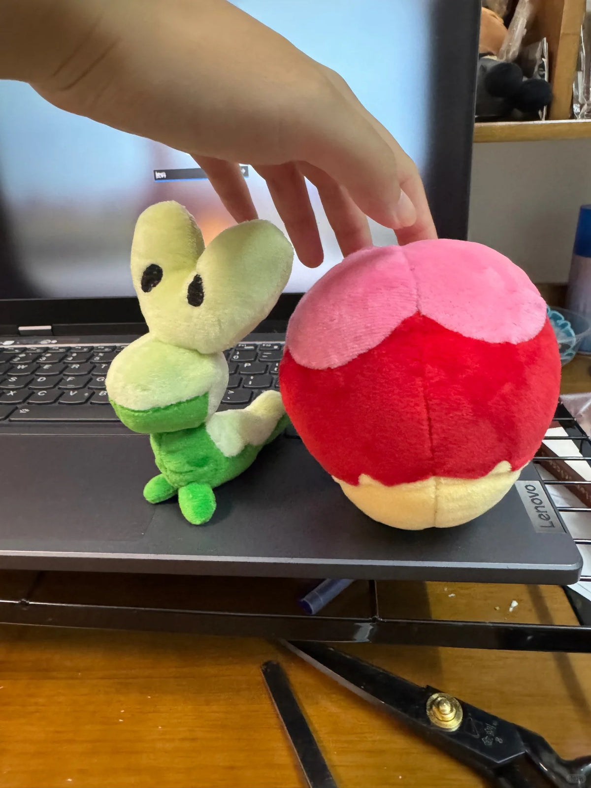 A Applin plush toy set on a laptop keyboard, featuring a green and cream frog-like character next to a red and pink strawberry plush, with a hand reaching toward the strawberry.