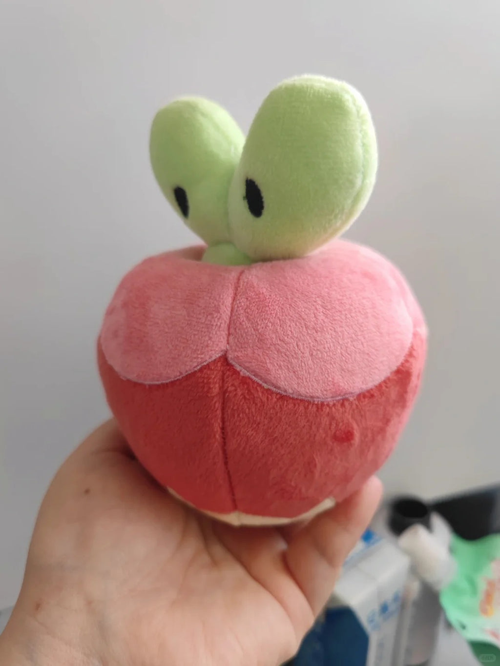 A Applin plush toy shaped like a stylized red apple with two light green leaf-like tops and small black eyes, being held in a person's hand against a plain indoor background.
