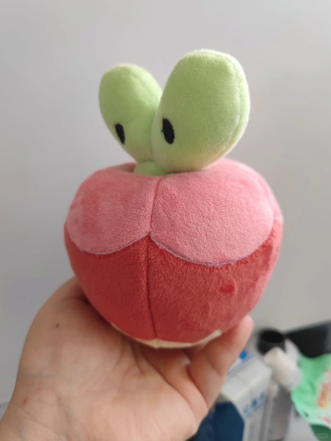 A Applin plush toy shaped like a stylized red apple with two light green leaf-like tops and small black eyes, being held in a person's hand against a plain indoor background.