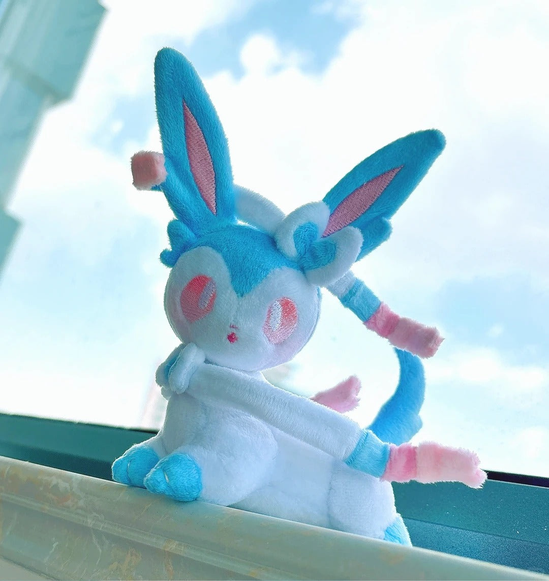 A plush toy of a blue and white Sylveon with pink accents, posed sitting on a windowsill with a bright sky background.