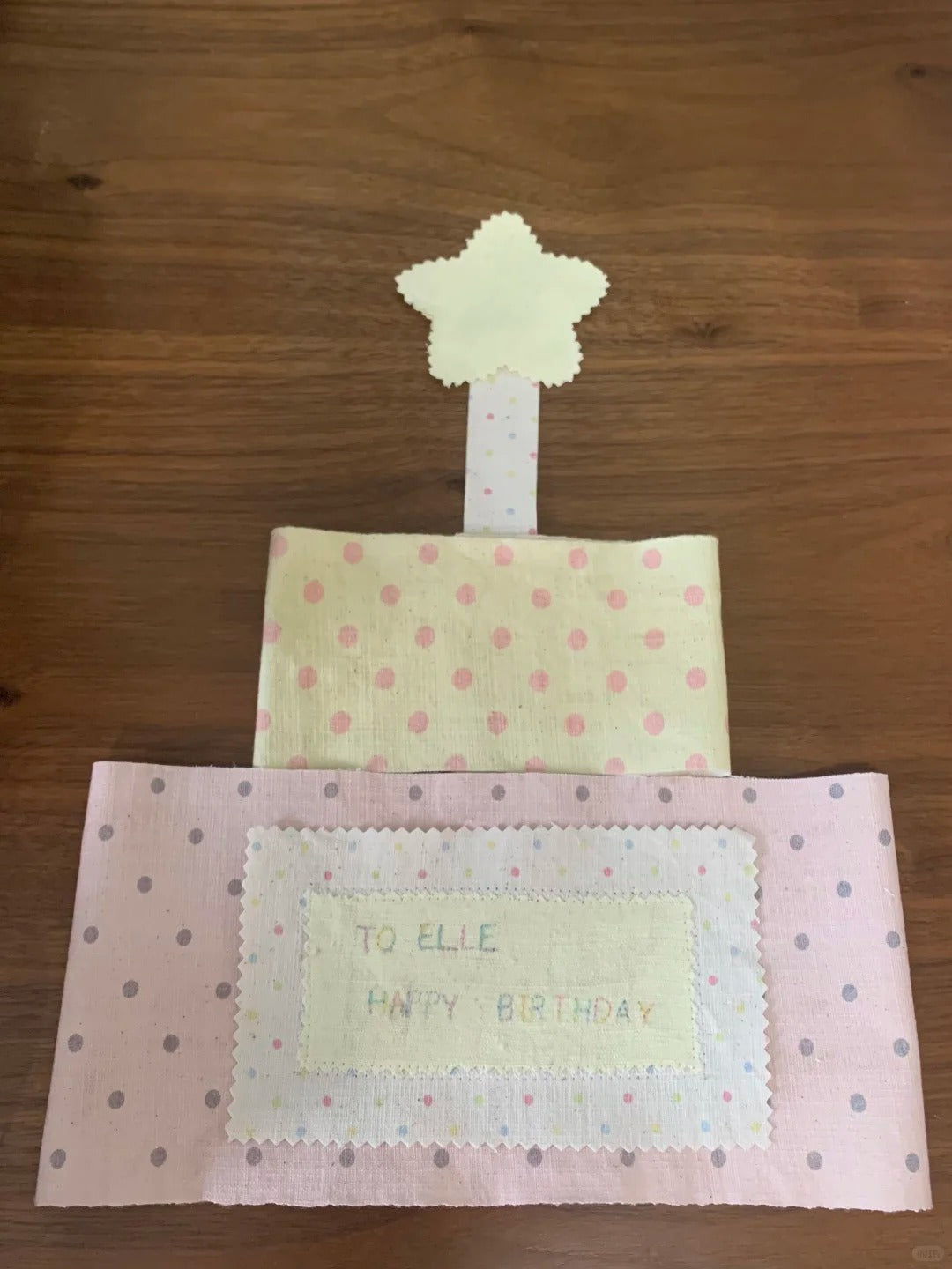 A handmade fabric birthday cake gift for a child named Elle, featuring two tiers with pink and gray polka dots, a cream star topper, and a central label that reads 'TO ELLE HAPPY BIRTHDAY'.