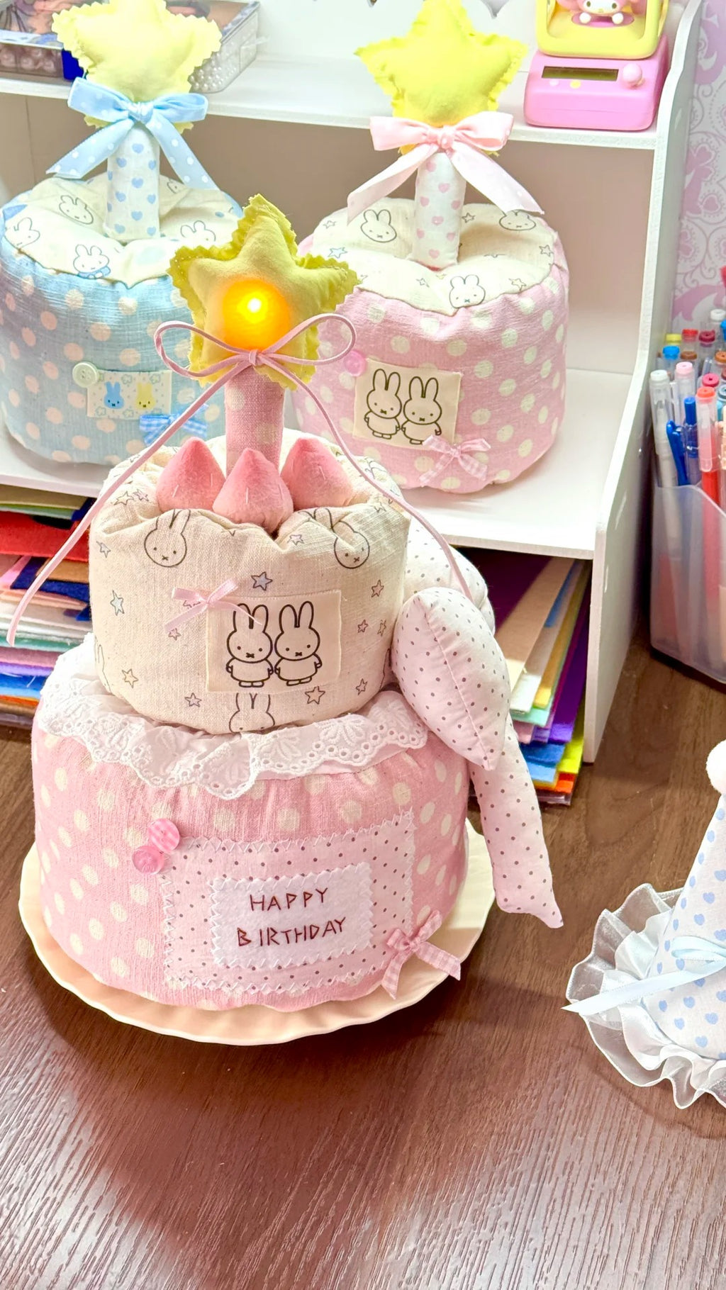 A pink and beige fabric cake-shaped birthday gift set with bunny illustrations, a star topper, and a 'HAPPY BIRTHDAY' label, displayed on a wooden surface with other similar items in the background.