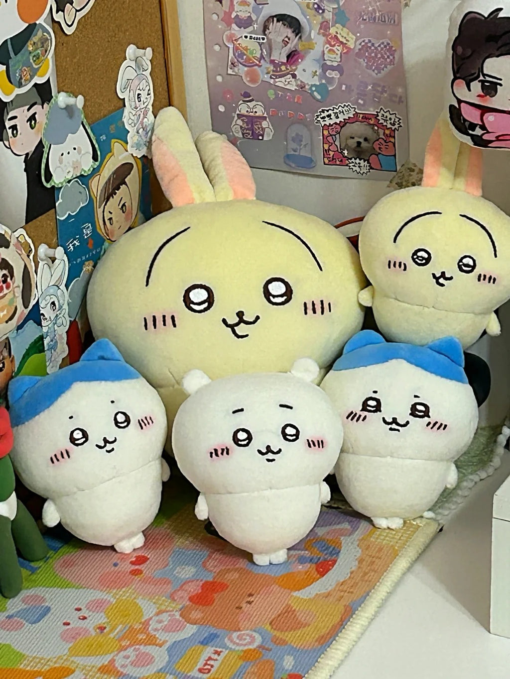 Five cute plush toys with chibi-style faces arranged together on a colorful play mat, with stickers and posters visible in the background.