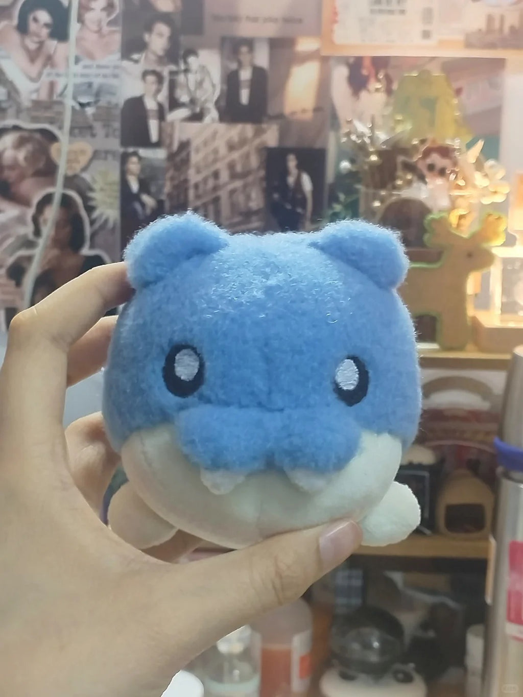 A hand holding a small blue Spheal plush toy with a shark-like or whale-like face, featuring black eyes and a white underside, against a busy indoor background with posters and shelves.