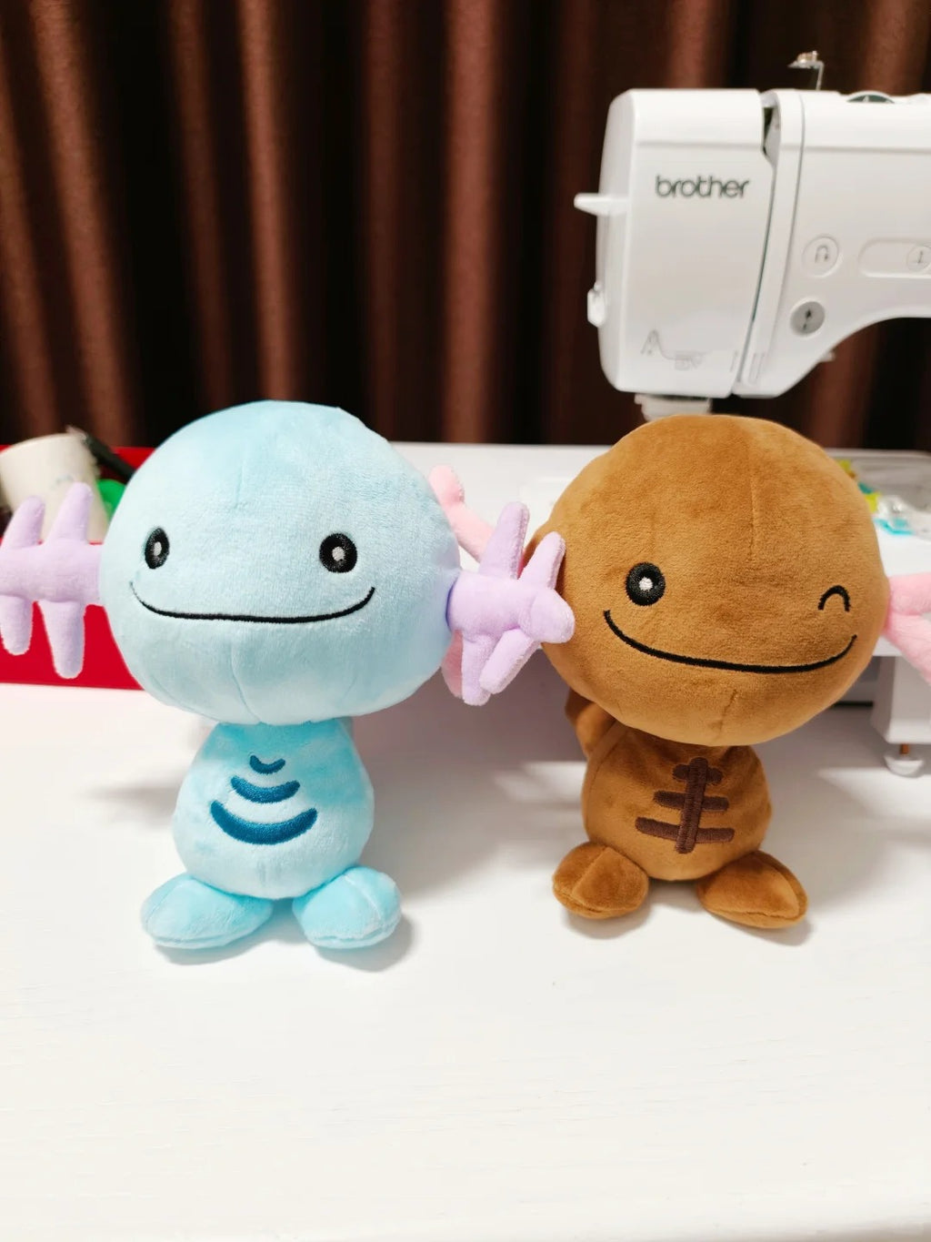 Two plush Wooper toy figures sitting on a white table in front of a sewing machine. The left figure is light blue with pink wing-like arms and a simple stitched smile.