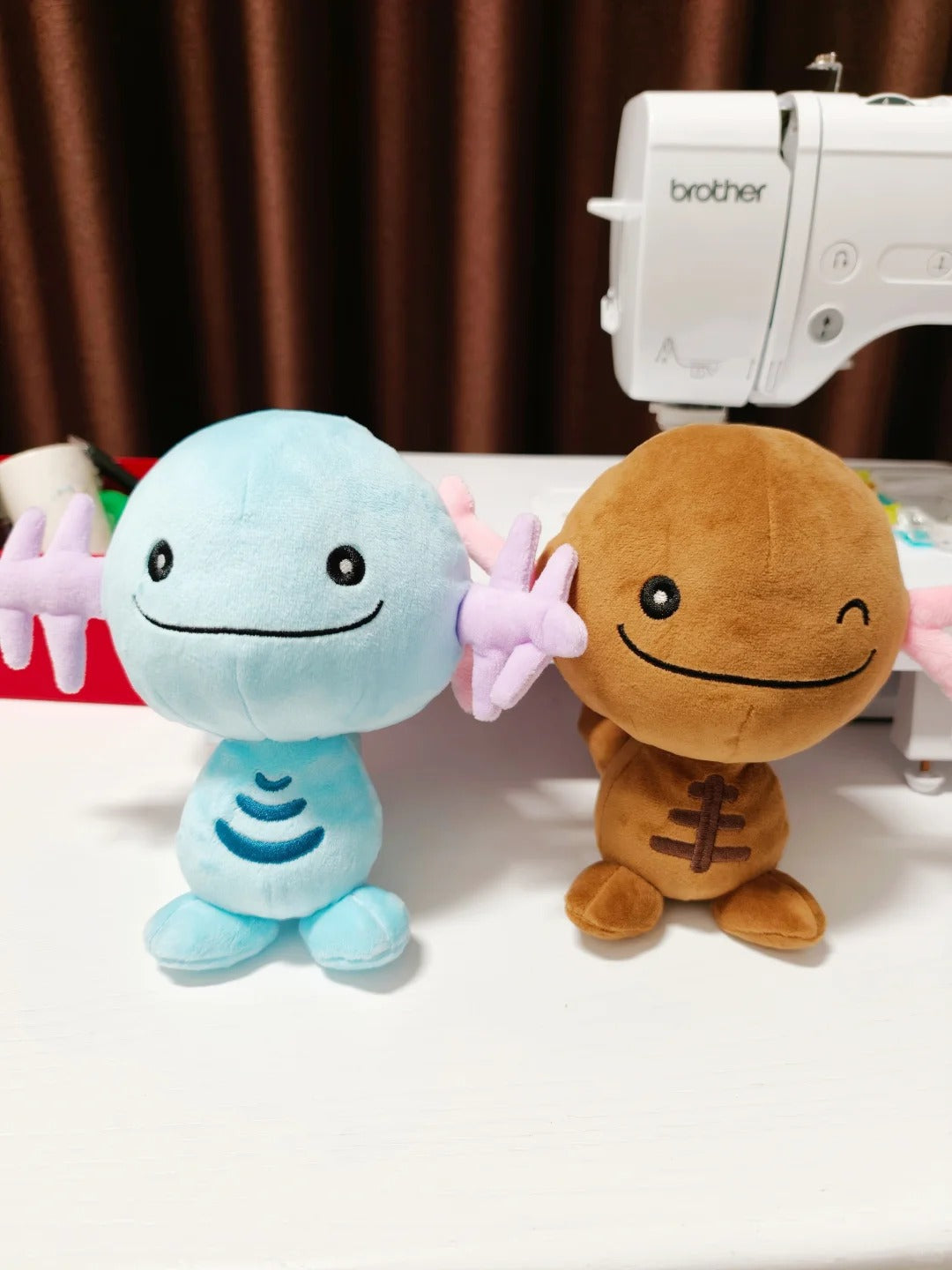 Two plush Wooper toy figures sitting on a white table in front of a sewing machine. The left figure is light blue with pink wing-like arms and a simple stitched smile.