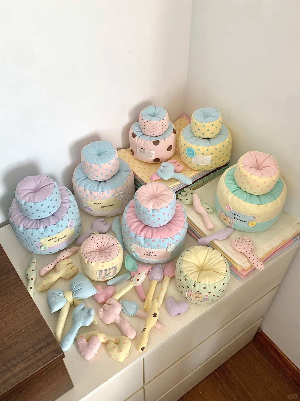A collection of pastel-colored baby keepsake boxes and small accessories arranged on a white dresser in a room corner. The boxes come in soft pastel shades of pink, blue, yellow, and green with polka dots, and are styled with small bows, ribbons, and plush toys.