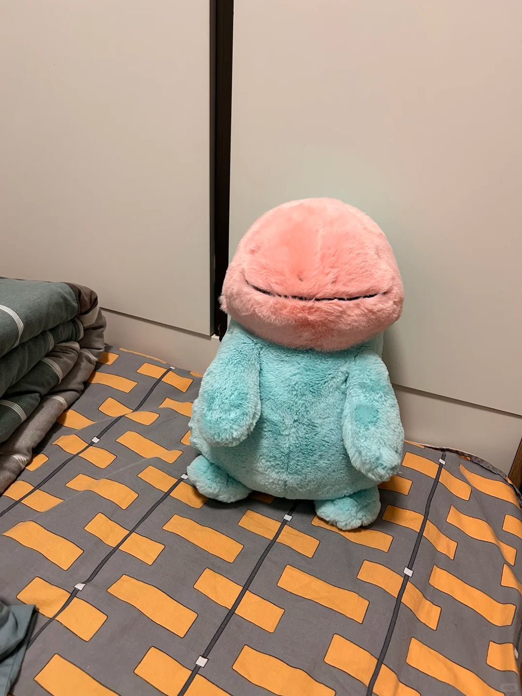 A  Quagsire plush toy resembling a cute blue creature with a pink dome-shaped head, sitting on a patterned gray and orange bedsheet in a bedroom.