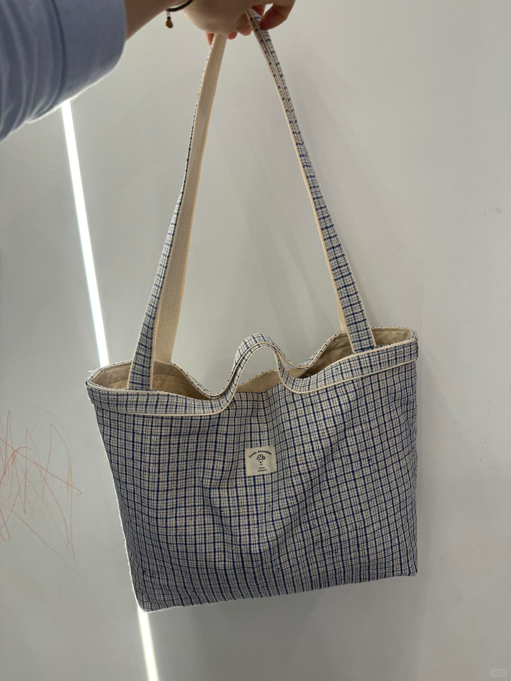 A canvas tote bag with a blue and white grid pattern and beige shoulder straps, held by a hand against a white wall.