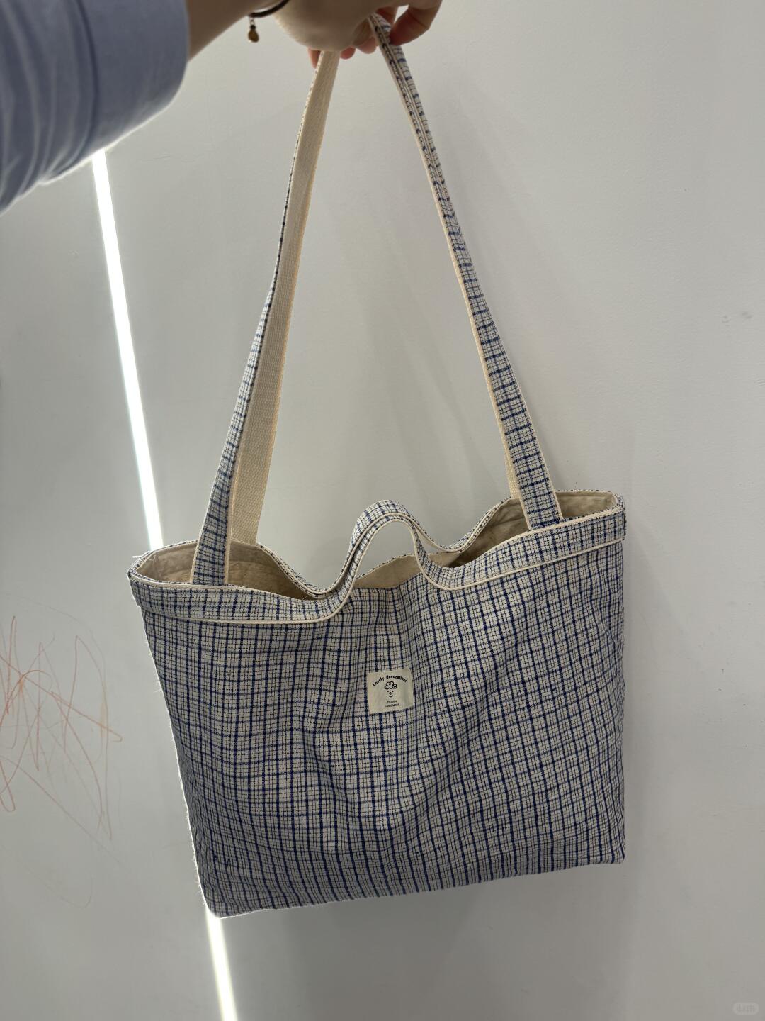 A canvas tote bag with a blue and white grid pattern and beige shoulder straps, held by a hand against a white wall.