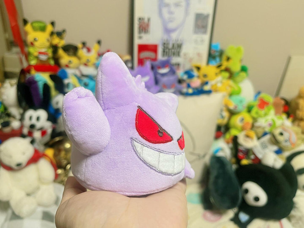 A small lavender Gengar plush toy resembling Gengar is held in a person's hand, with a collection of colorful plush toys and posters blurred in the background.