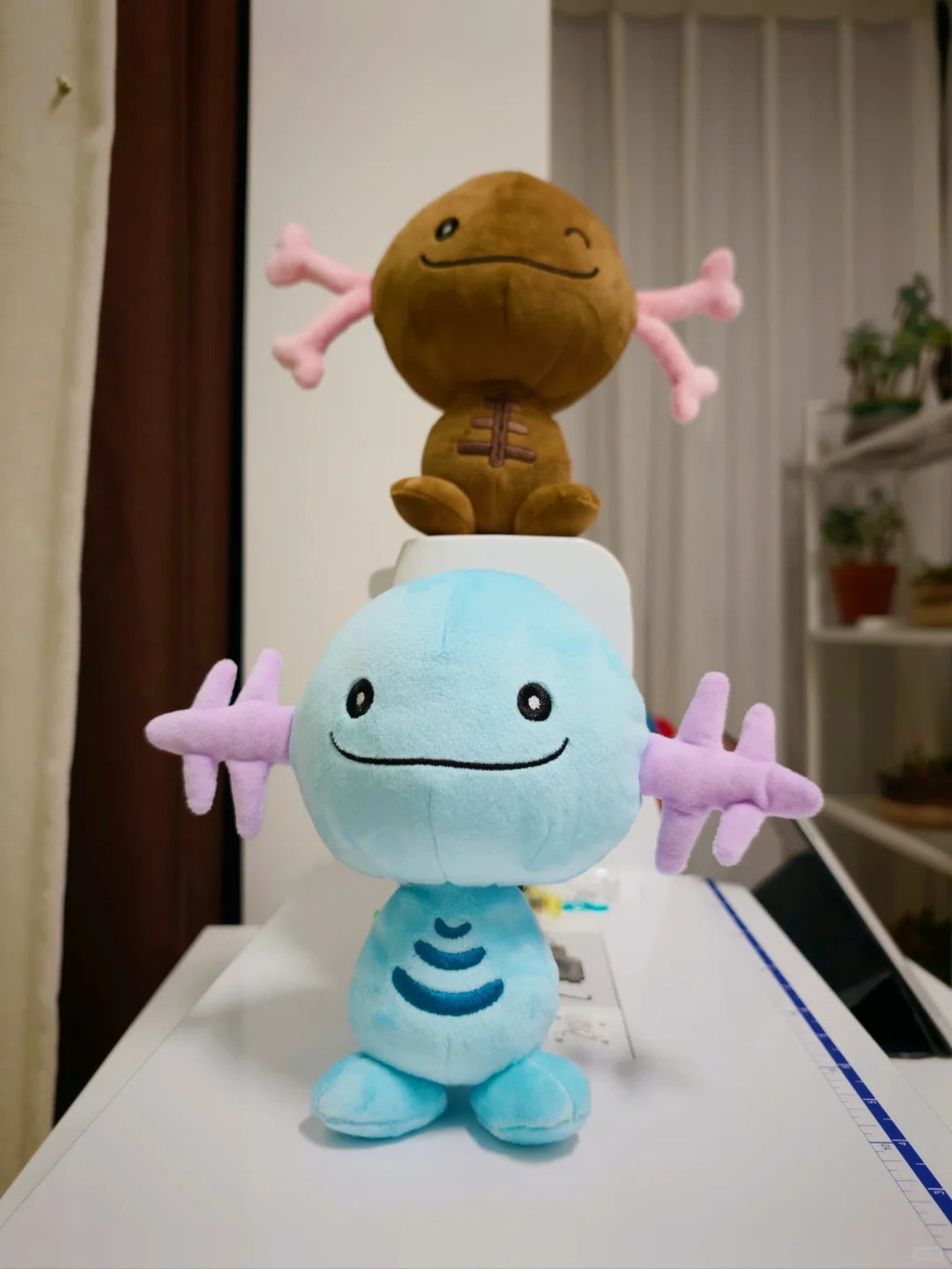 Two plush Wooper toys arranged on a white surface in an indoor room. The front plush is light blue with pink star-like arms and a stitched smile.