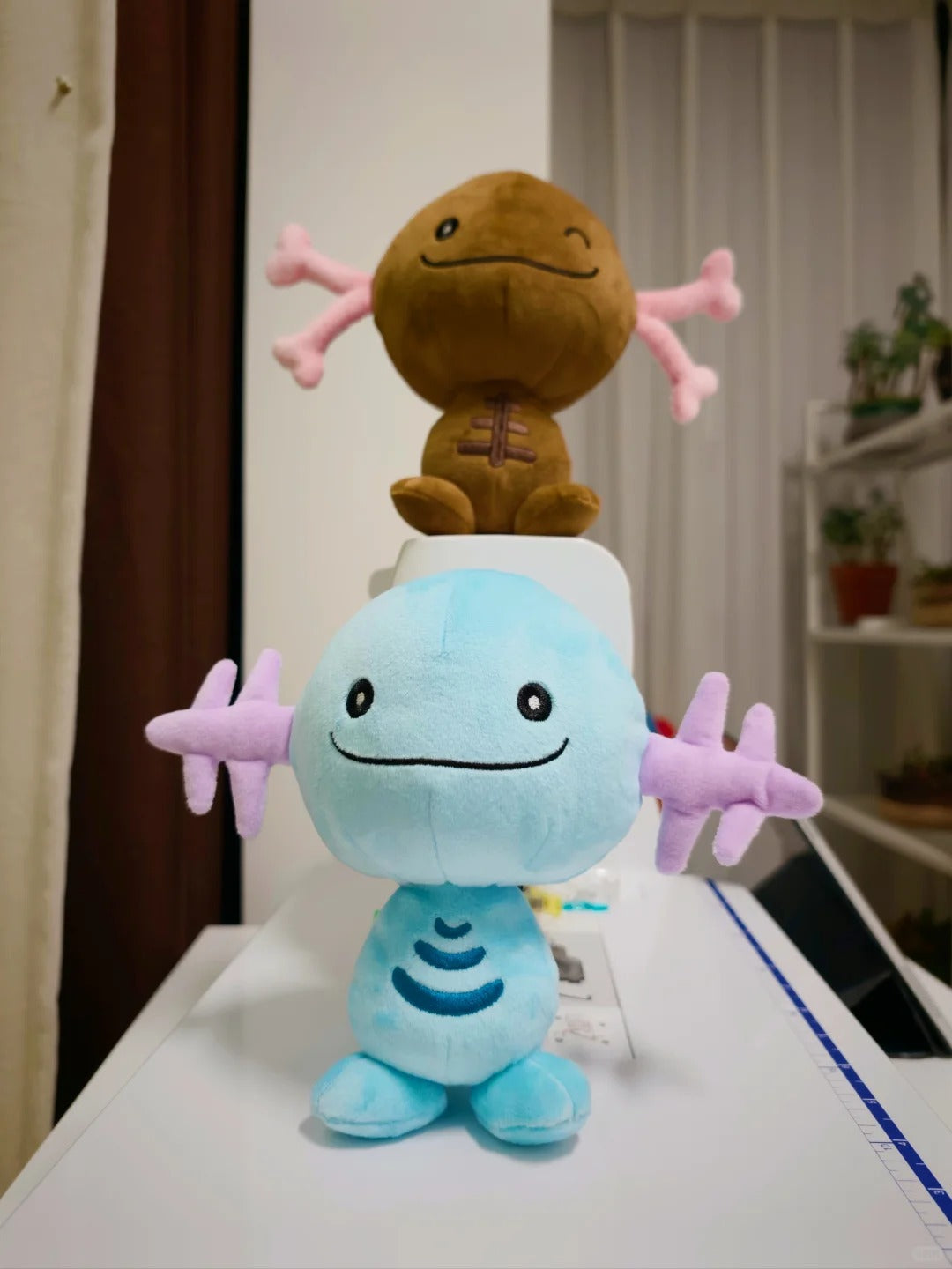 Two plush Wooper toys arranged on a white surface in an indoor room. The front plush is light blue with pink star-like arms and a stitched smile.