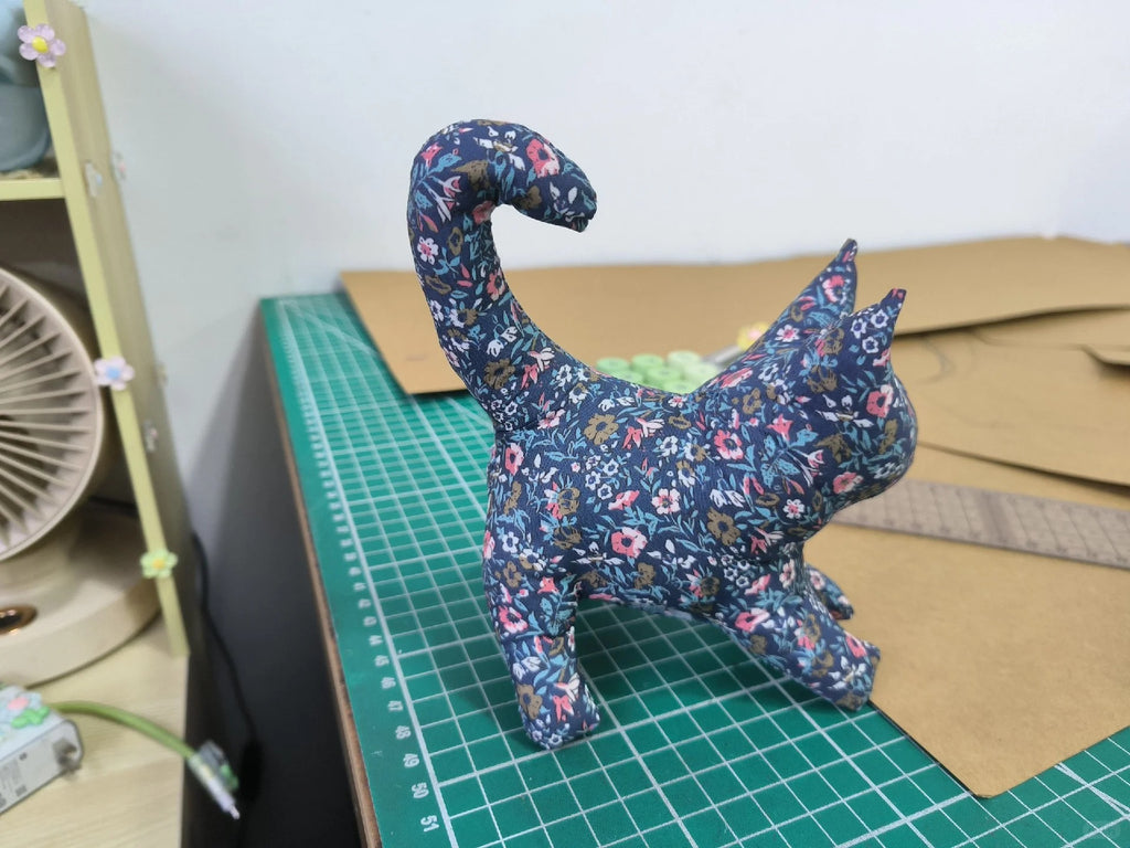 A small fabric cat figurine with a blue floral print stands on a green cutting mat on a workbench.